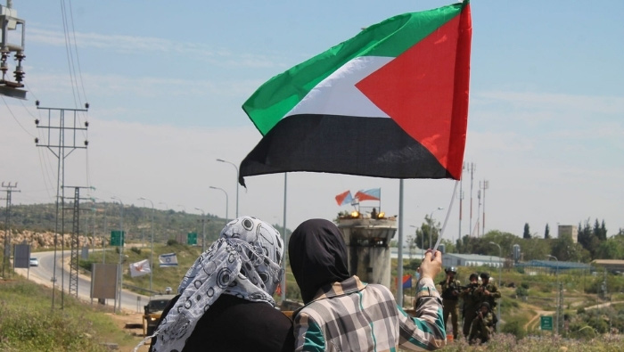 Two palestinian women waving the palestinina flan in the direction of an Israeli roadblock